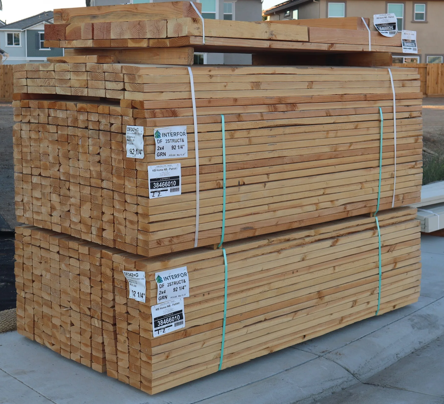 Two banded bundles of sawn framing lumber with mill labels, set down at the curb, houses under construction behind them.