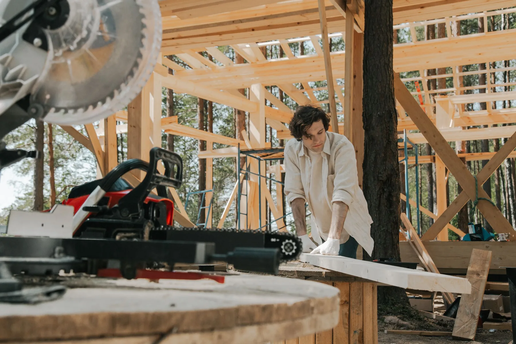 A carpenter guides a board into a mitre saw mounted on a stand inside a timber frame, with cut lengths and trees behind him.