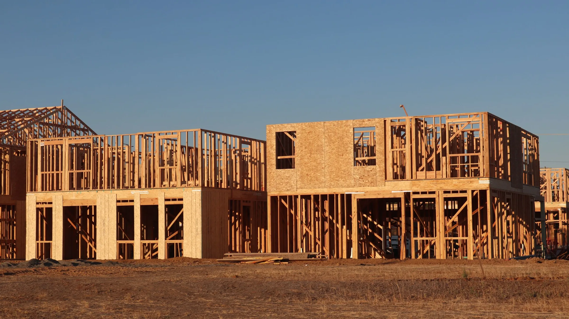 A row of two-story houses at the framing stage, part of the walls already closed with OSB sheathing, under a cloudless sky.