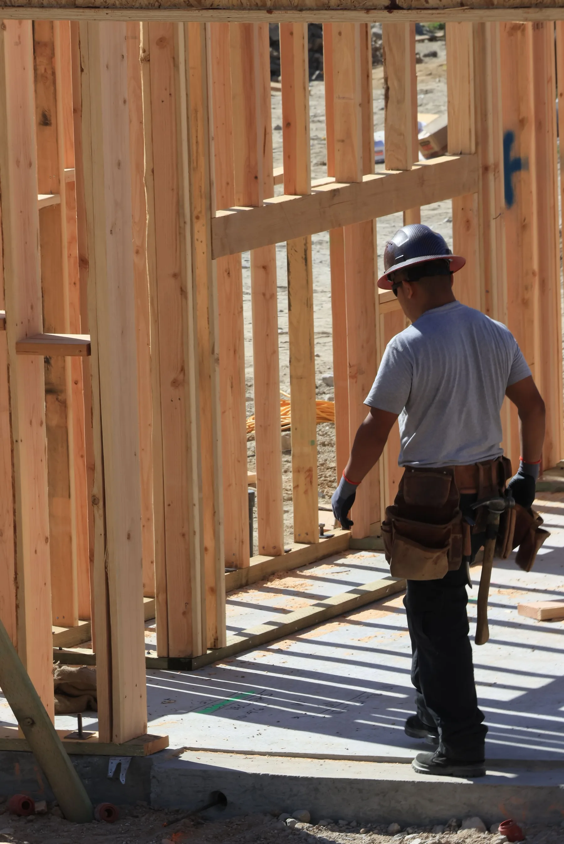 A worker in a hard hat and a leather tool belt steps through a door opening in a newly raised stud wall standing on a concrete slab.