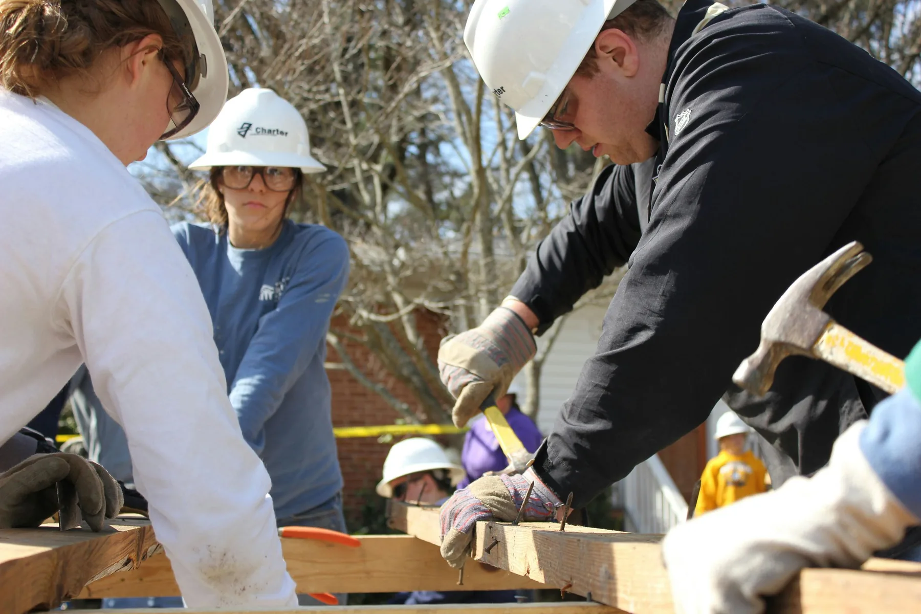 Several people in hard hats and work gloves hold a timber wall frame down on the ground while one of them drives a nail with a hammer.