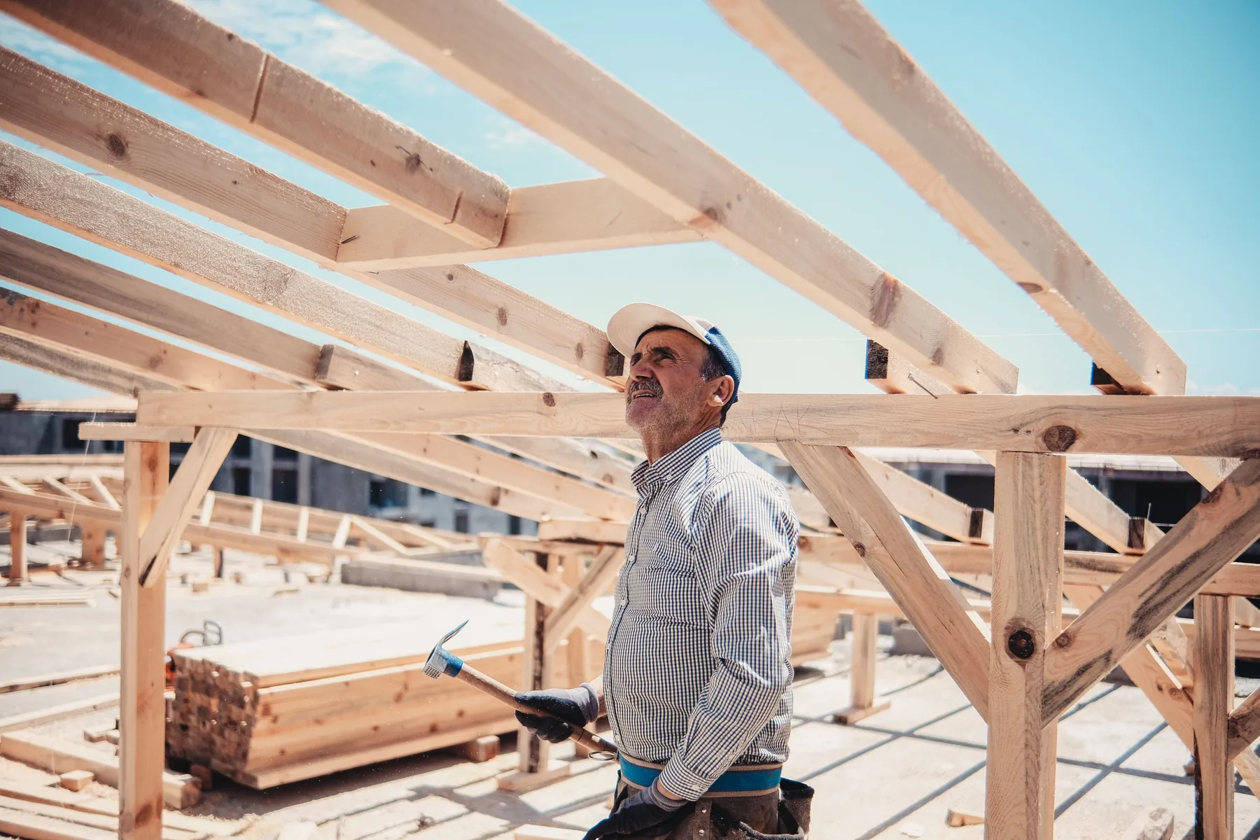 A worker with a hammer in his hand looks up at freshly erected rafters and beams of a roof frame against a bright sky.