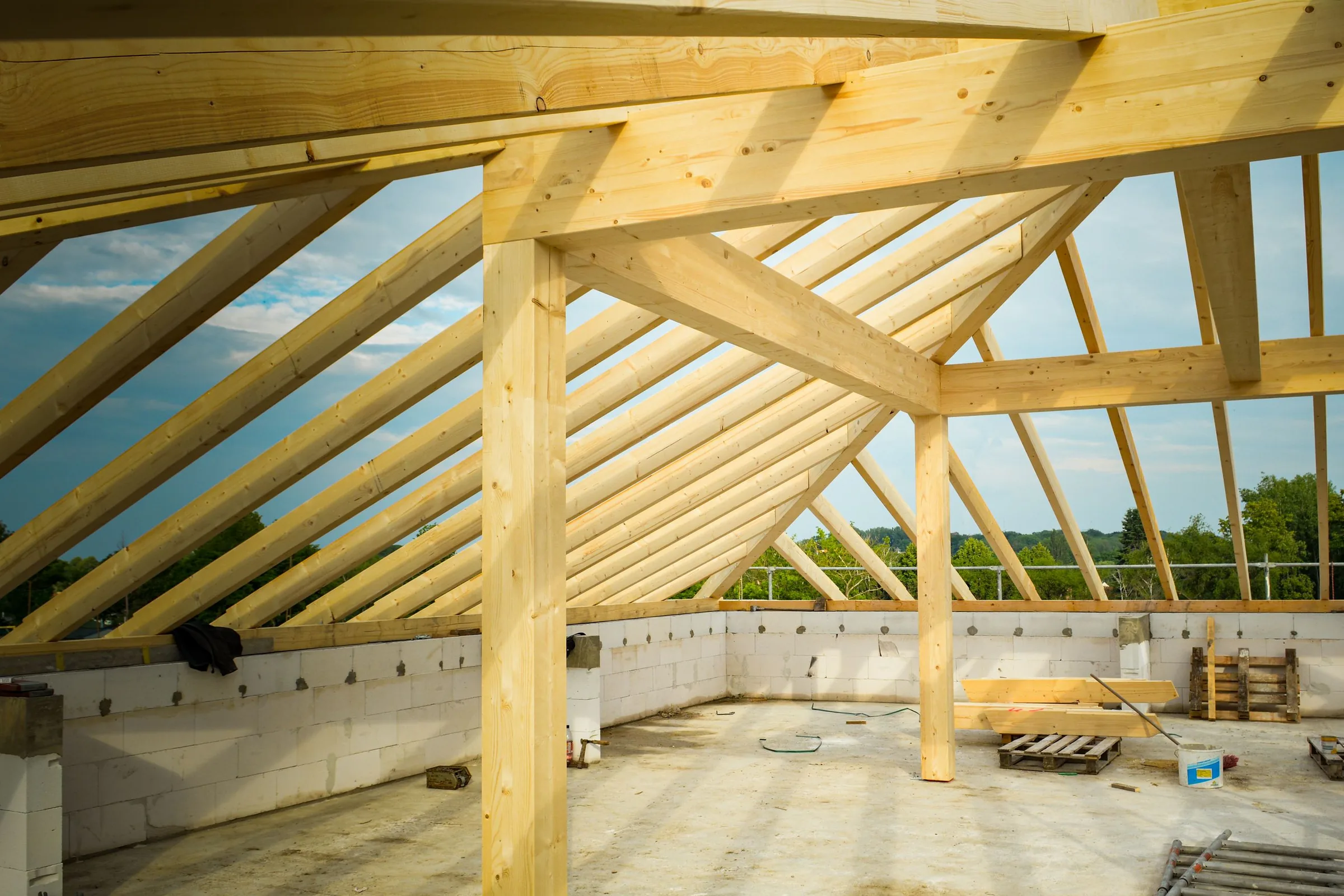 A heavy timber roof structure seen from below: rafters, purlins, a ridge beam and posts standing against open sky on top of masonry walls.
