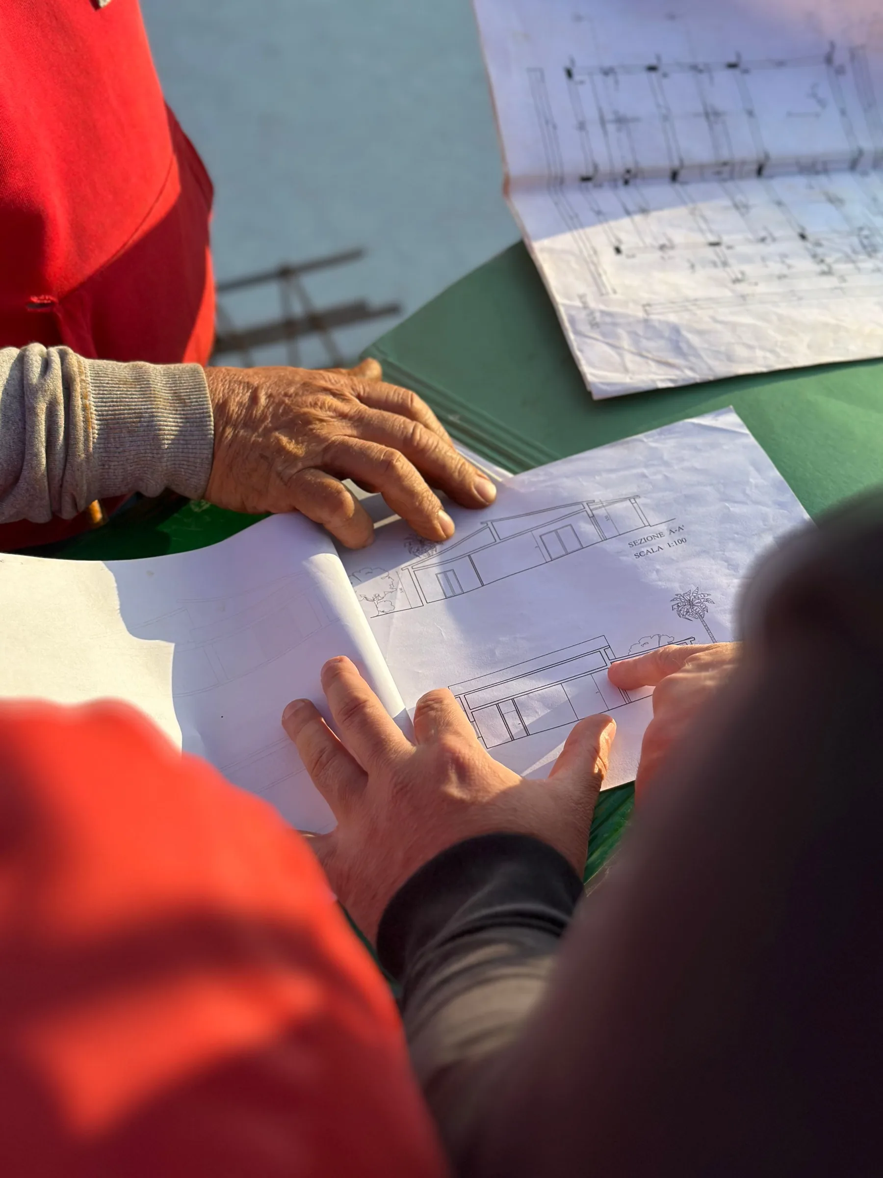 Two people in orange high-visibility clothing lean over a table outdoors, their hands spread across construction drawings and a folder.