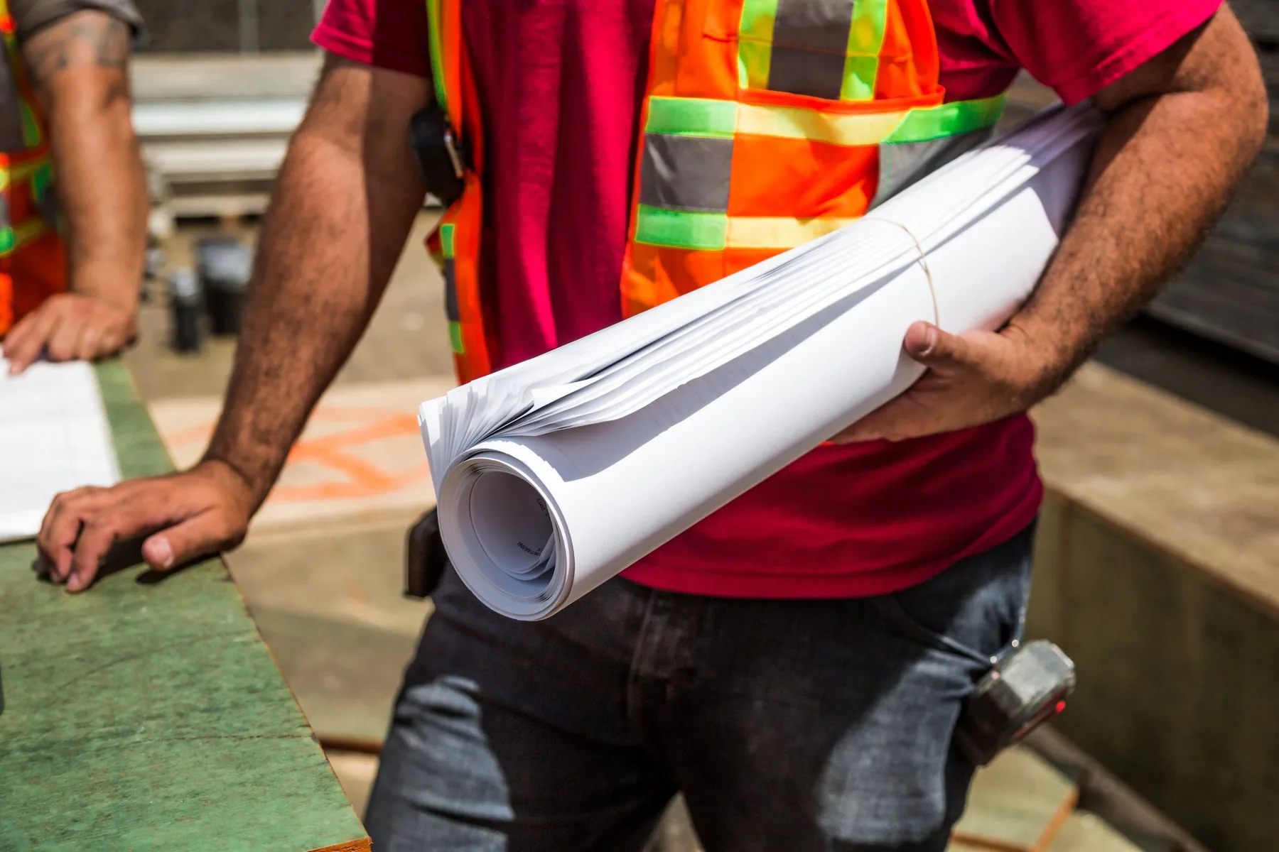 A worker in a high-visibility vest carries a rolled set of drawings under one arm, a hammer hanging from the belt, on a construction site.