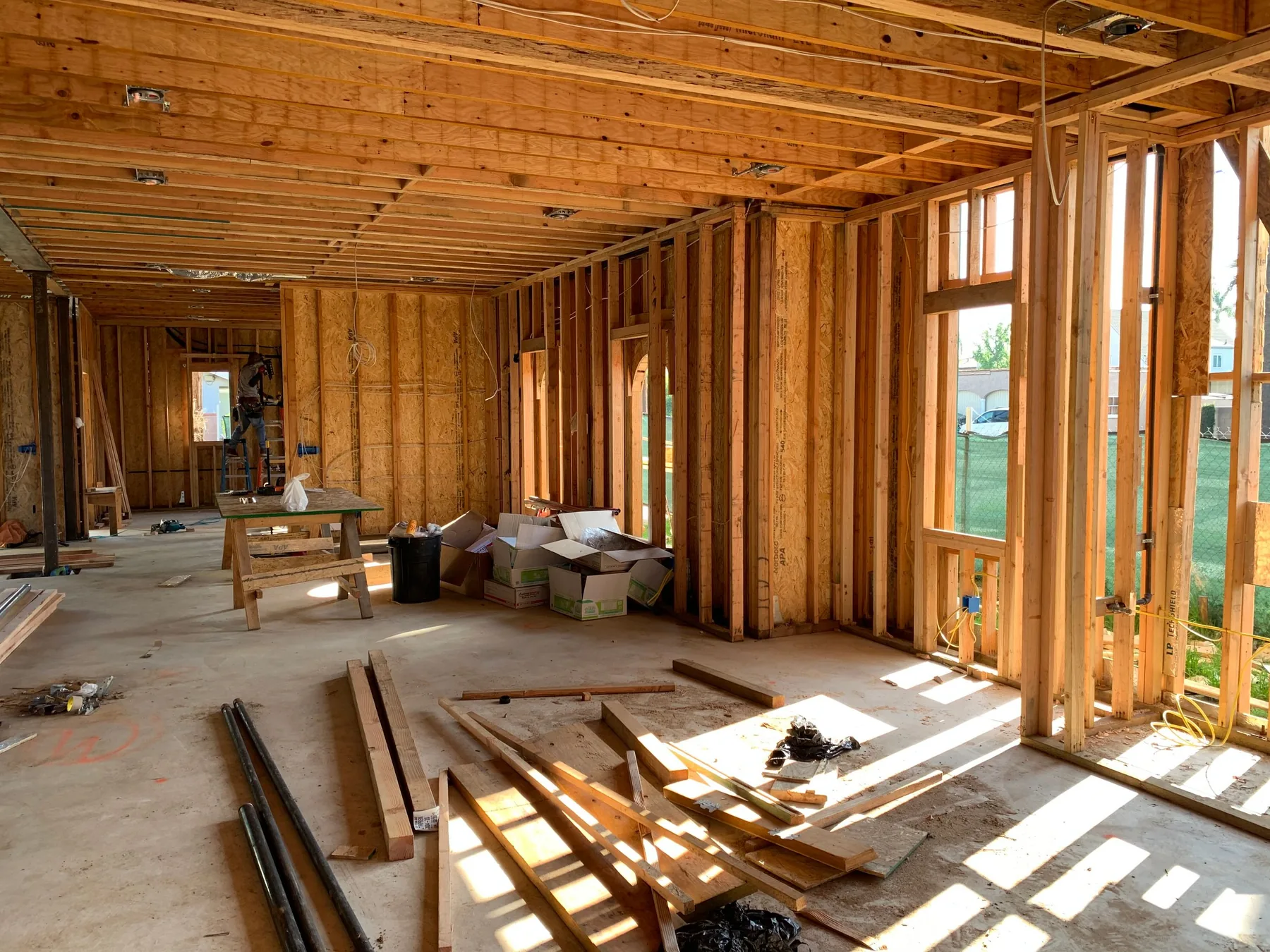 Low sunlight falls through the openings of a house shell onto stud walls, ceiling joists, offcuts and packs of material lying on the subfloor.