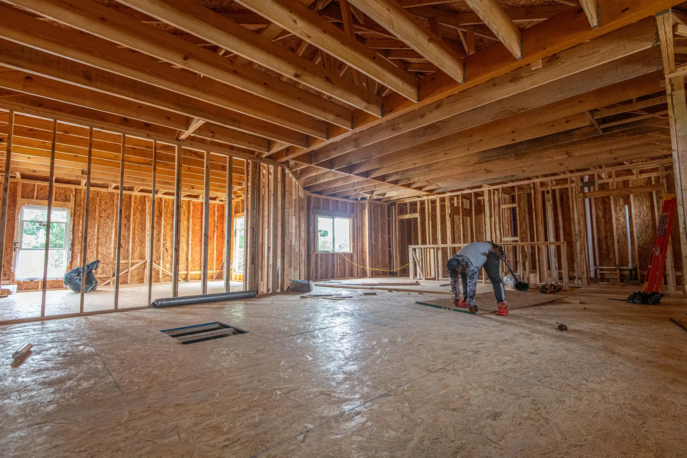 The inside of a house shell: stud walls, ceiling joists, open window holes and a plywood subfloor, with one worker crouched at the far end.